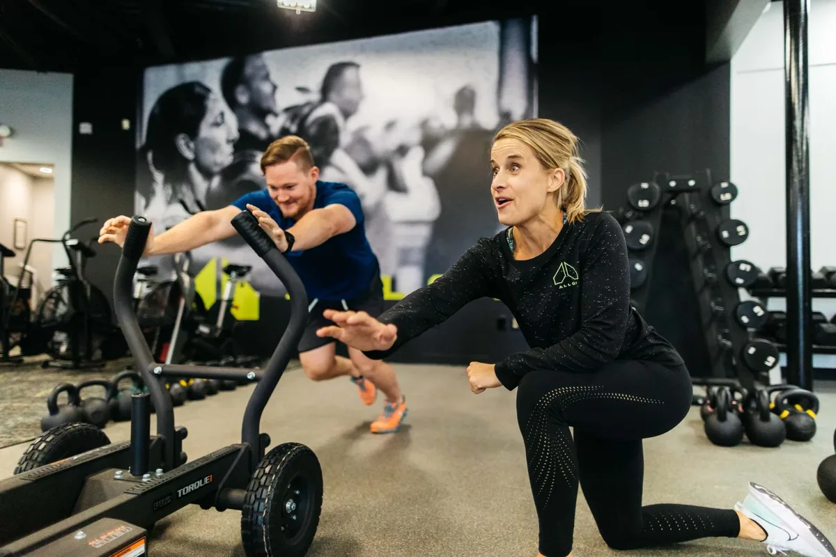 A member pushes a weighted sled across the studio floor while a coach encourages him from the side