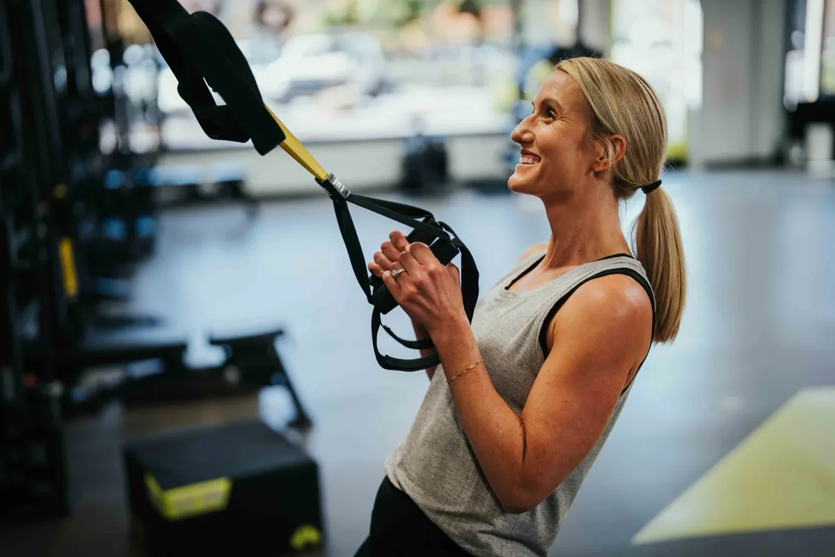 A smiling member leans back into a TRX row during a coached session at an Alloy studio