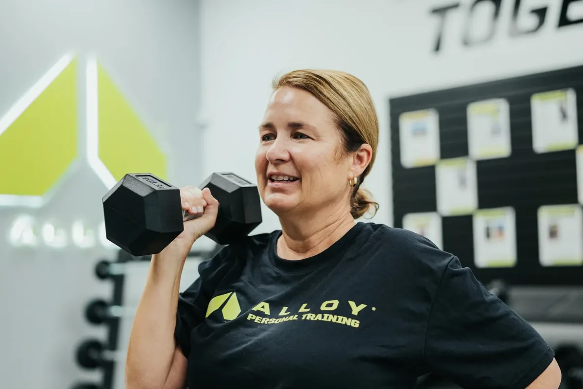 A member presses a dumbbell at shoulder height during a coached session at an Alloy studio