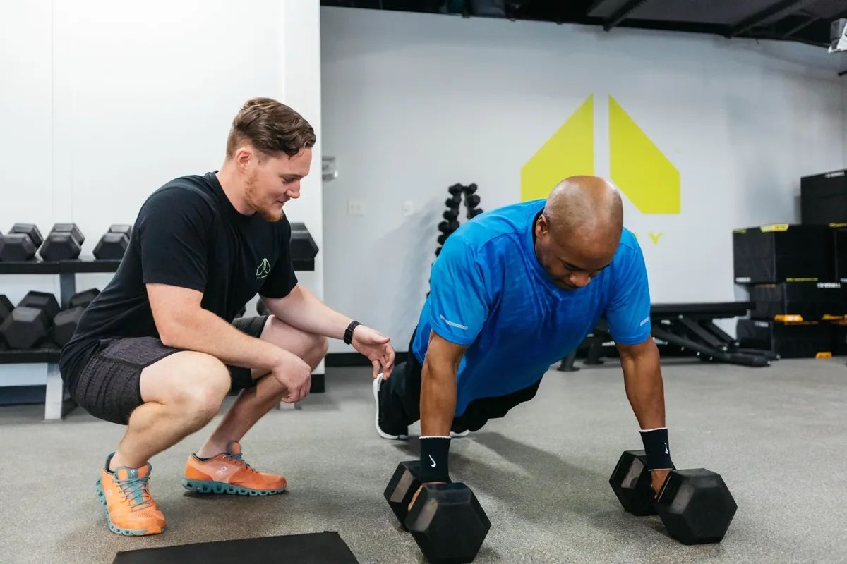 A coach kneels to watch a member's form during renegade rows at an Alloy Personal Training studio
