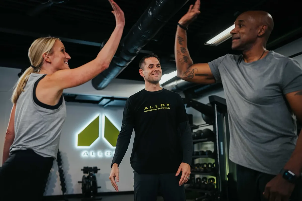 Two members and a coach share high fives during a small group session at an Alloy Personal Training studio