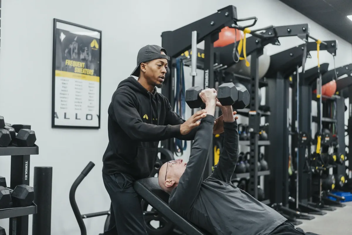 A coach guides an older member's dumbbell press from behind the bench
