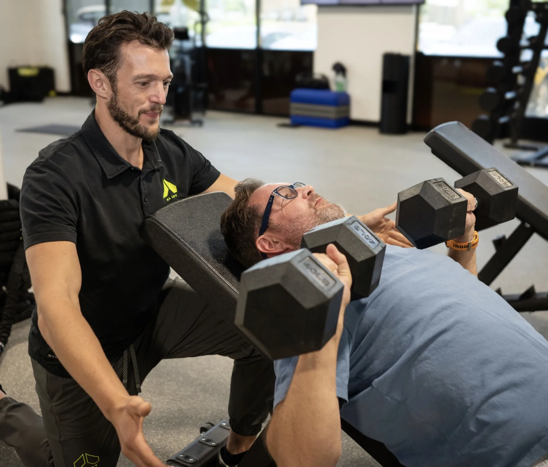 A trainer leans in to spot a member pressing dumbbells on a bench, one on one