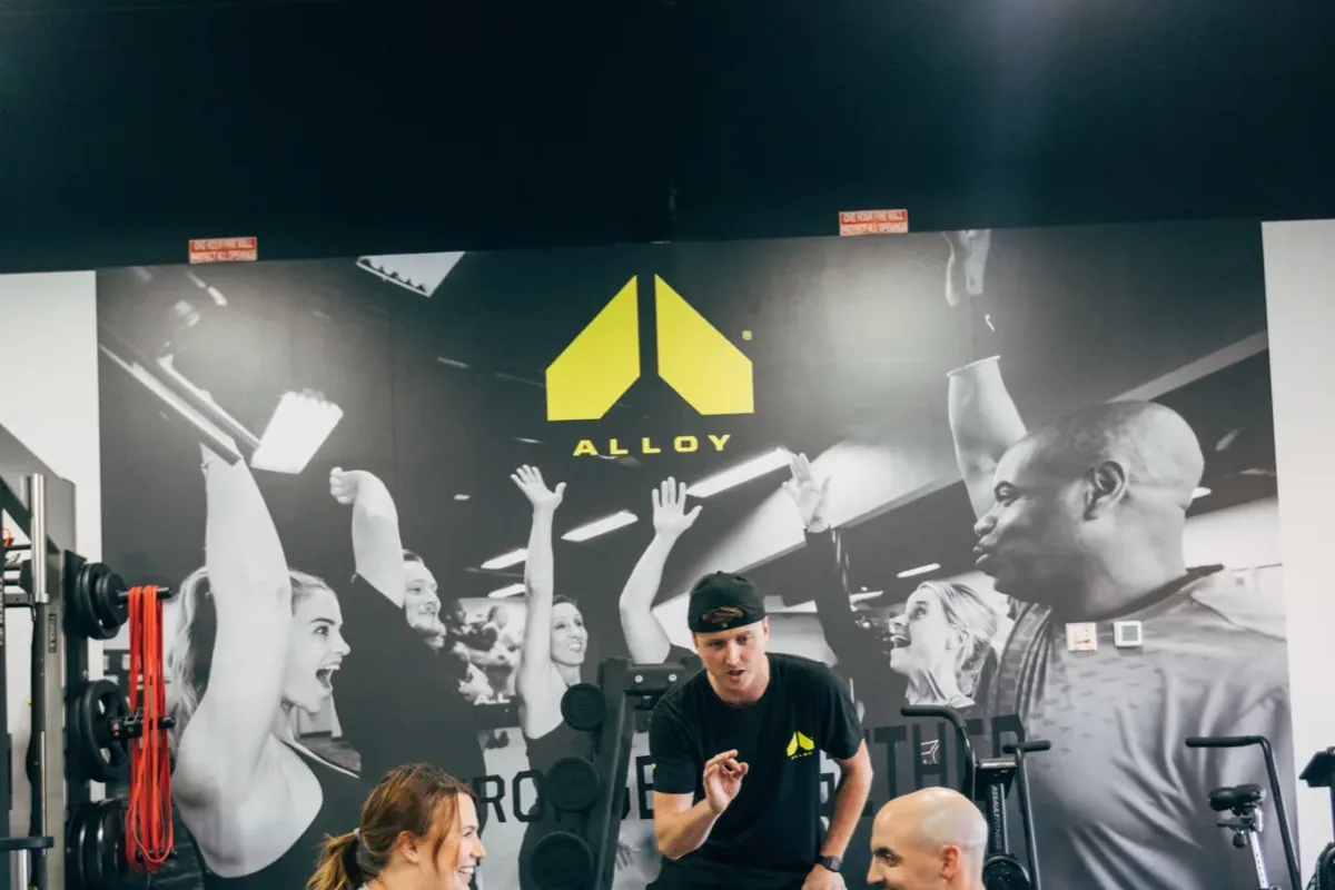 A coach talks two members through the next exercise beneath the Stronger Together mural at an Alloy Personal Training studio