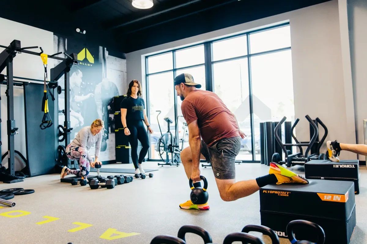 A coach leads a small group of adults training with kettlebells and dumbbells at an Alloy Personal Training studio