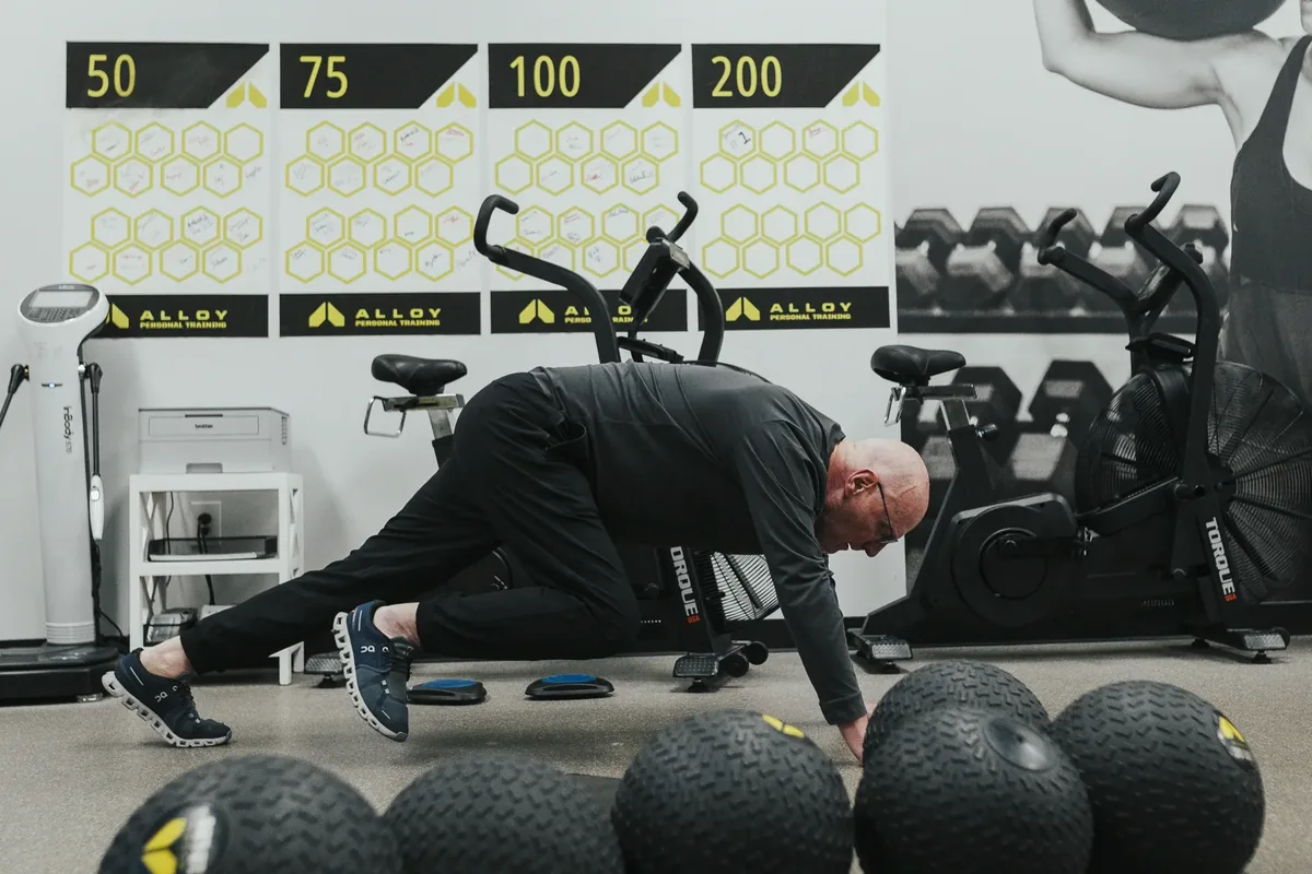 An older member works through a floor mobility drill near the member milestone wall at an Alloy Personal Training studio