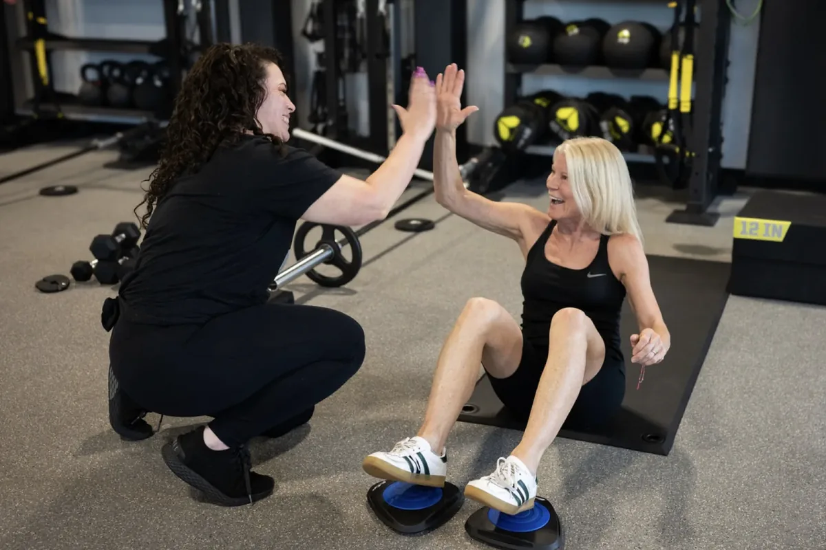 A coach and a smiling member share a high five during a core exercise on glider discs