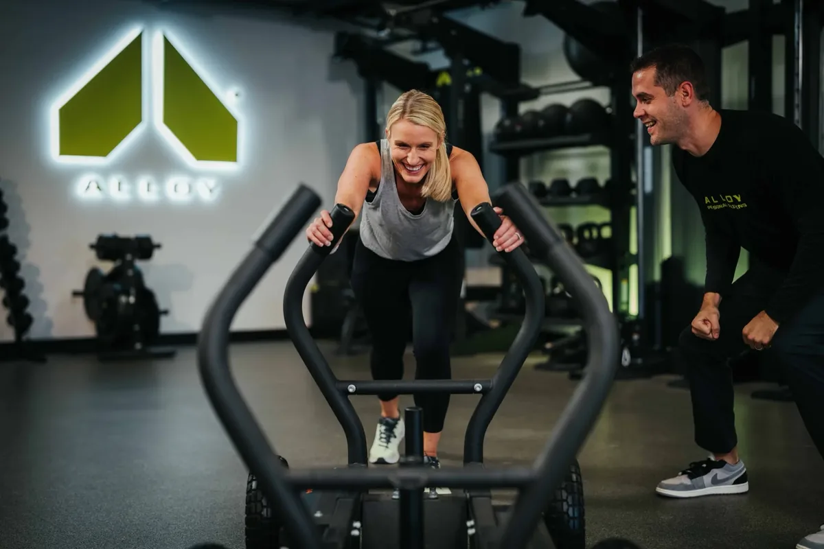 A coach encourages a smiling member as she pushes a training sled across the studio floor