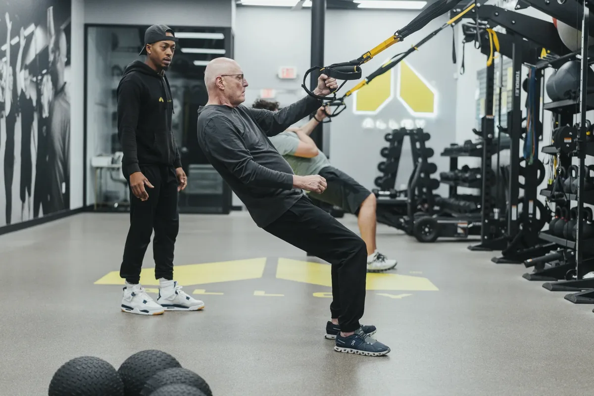 A member leans back into TRX straps under a coach's eye while another member trains in the background at an Alloy studio