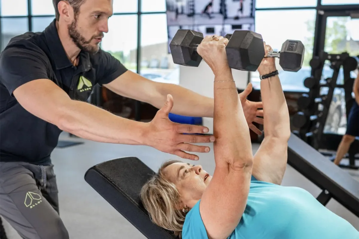 A coach guides an older woman's arm position as she presses dumbbells on an incline bench