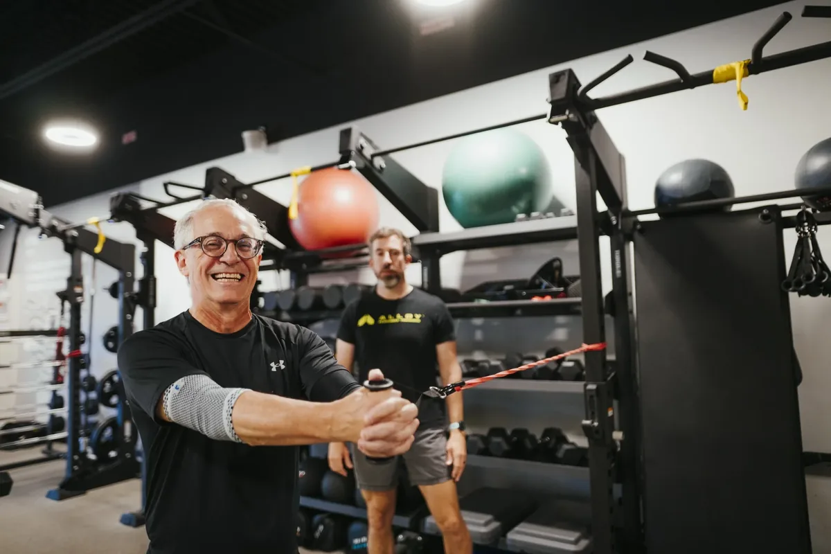 An older member performs a banded cable exercise while his coach supervises behind him